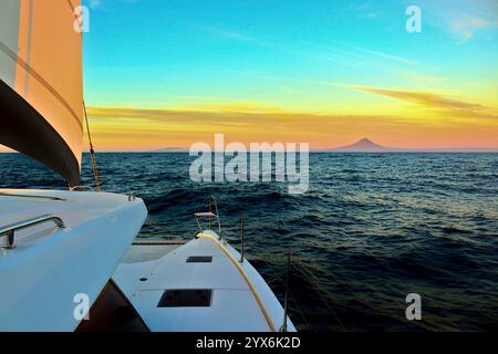 Una barca a vela scivola nel tramonto con la silhouette di Faial Island all'orizzonte, incorniciata dal bagliore dorato del vasto Oceano Atlantico. Foto Stock