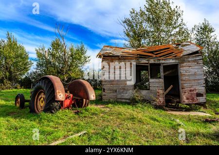 Un vecchio casale in rovina si trova in un campo erboso, con un trattore rosso arrugginito parcheggiato di fronte. La casa sembra essere in uno stato di disrepa Foto Stock