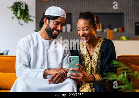 Felice bella giovane coppia araba con abiti emirati insieme a casa utilizzando il cellulare per lo shopping online - uomo e donna mediorientale con mestieri Foto Stock
