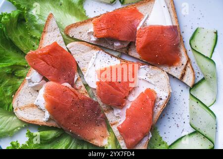 Panini con formaggio fresco e fette di trota salata su un piatto bianco con cetriolo ed erbe aromatiche. Foto di alta qualità Foto Stock