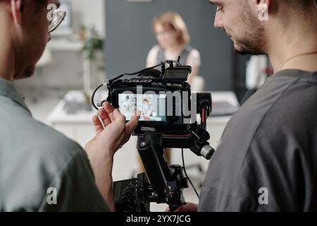Primo piano medio di videografi irriconoscibili che guardano l'esposizione della fotocamera e discutono la scena con il modello seduto al tavolo sullo sfondo Foto Stock