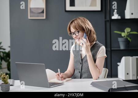 Medio primo piano di una giovane elegante donna caucasica seduta alla scrivania che parla sullo smartphone con il cliente e prende appunti Foto Stock