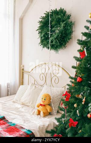 L'accogliente camera da letto presenta un albero di Natale splendidamente decorato con ornamenti e peluche di pan di zenzero accanto ai cuscini. Letto con biancheria bianca e cushi decorativo Foto Stock