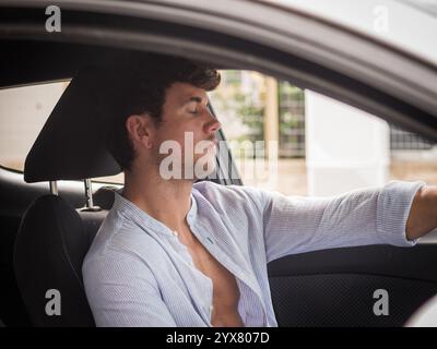 Un giovane uomo si rilassa con gli occhi chiusi mentre è seduto al posto di guida di un'auto parcheggiata. Sembra calmo e tranquillo in un ambiente informale durante il giorno. Foto Stock