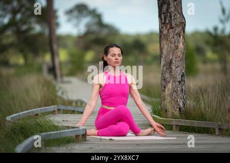 Donna sportiva seduta su una passerella a meditare e praticare yoga indossando abbigliamento sportivo rosa Foto Stock
