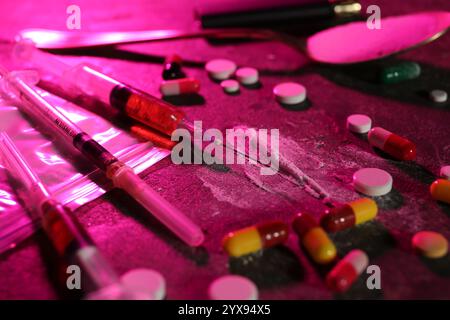 Tossicodipendenza. Sacchetto di plastica con polvere, siringhe e pillole sul tavolo in luce colorata, primo piano Foto Stock