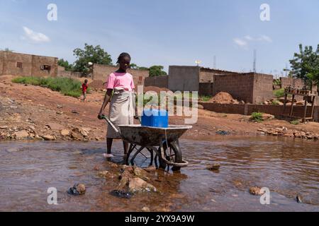 Scena rurale di una giovane bambina africana che attraversa un fiume con la sua pesante carriola che trasporta una tanica d'acqua. Foto Stock