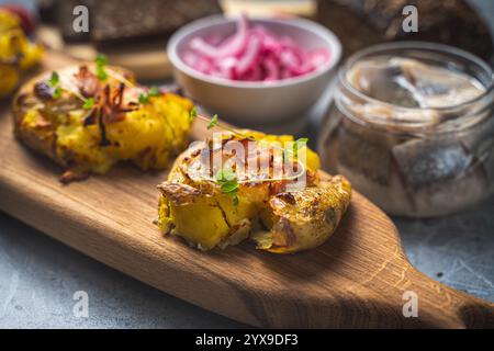 Patate al forno con formaggio e pancetta schiacciate su un asse di legno su un tavolo grigio con un barattolo di aringhe, cipolle rosse sottaceto e pane di segale. Alta qualità p Foto Stock