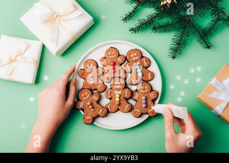 Le mani di una ragazza decorano biscotti di pan di zenzero natalizi su un piatto bianco circondato da regali avvolti ed elementi festivi su uno sfondo verde. Vie superiori Foto Stock