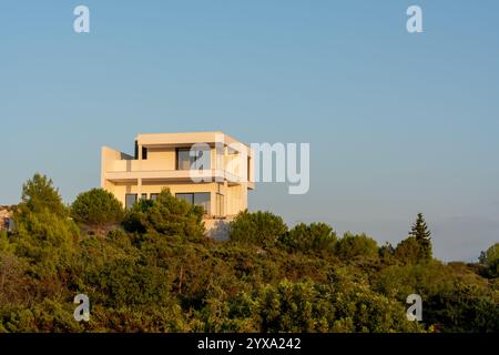 Moderna villa bianca con grandi finestre in vetro circondata da lussureggianti alberi verdi sulla cima della collina durante l'ora d'oro. Concetto di immobili di lusso, contemporaneo Foto Stock