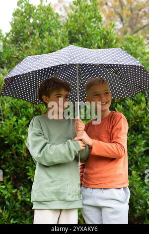 Due ragazzi che condividono un ombrello a pois, sorridendo felicemente in un giardino verde Foto Stock