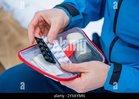 Primo piano delle mani di un alpinista che apre un kit di pronto soccorso contenente pillole, bende e altre forniture mediche mentre si trova in un ambiente di montagna innevato durante l'inverno Foto Stock