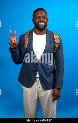 Giovane imprenditore nero che mostra due dita indossando vestito e zaino, isolato su sfondo blu studio Foto Stock