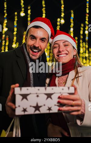 Verticale. Momento natalizio notturno: Amanti sorridenti che reggono un regalo rosso festoso, circondati da luci incantevoli, incarnano l'emozione pura e il romanticismo di coppia della stagione natalizia di dicembre. Foto di alta qualità Foto Stock