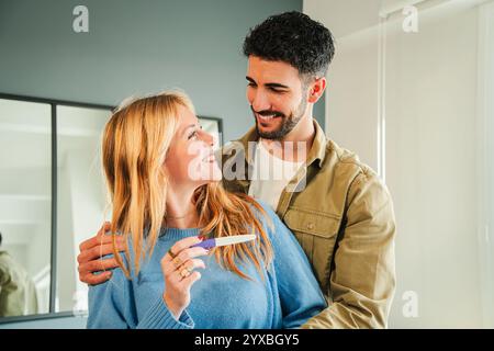 Coppia eccitata che celebra insieme le notizie della genitorialità a casa, sorridendo e tenendo un test di gravidanza positivo, condividendo momenti gioiosi ed emotivi mentre abbracciano, esprimendo felicità e anticipazione. Foto di alta qualità Foto Stock