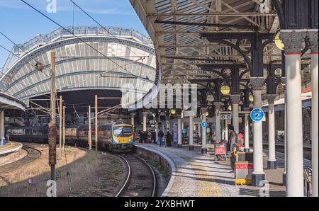 Un treno arriva a una fermata presso la piattaforma della stazione ferroviaria. Uno storico arco di ferro copre l'ingresso della stazione e i passeggeri aspettano di salire a bordo del treno Foto Stock