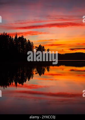 Le vivaci tonalità di arancione, rosa e viola riempiono il cielo al tramonto, mentre il sole tramonta dietro sagome scure di alberi. L'acqua serena rispecchia questo respiro Foto Stock