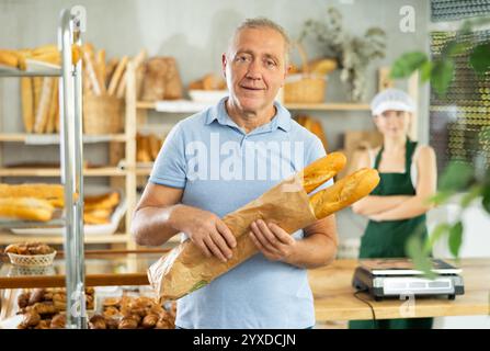 L'acquirente europeo di un uomo anziano si trova con orgoglio di fronte al pane fresco che tiene le baguette appena sfornate nel negozio di panetteria Foto Stock