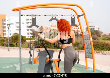 Donna in forma che si allena con una macchina stepper nella palestra all'aperto. La vista posteriore della femmina rossa in abbigliamento sportivo si esercita al parco durante il giorno. Foto Stock