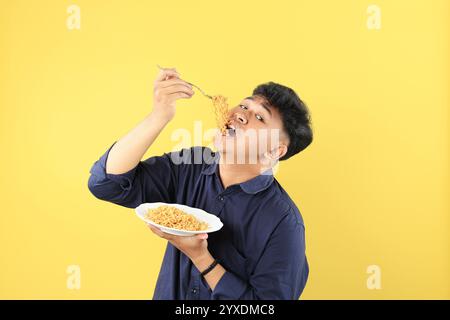 Felice ragazzo asiatico che indossa camicia blu mangia deliziosa mie Goreng su piatto bianco usando forchetta. Isolato su sfondo giallo Foto Stock