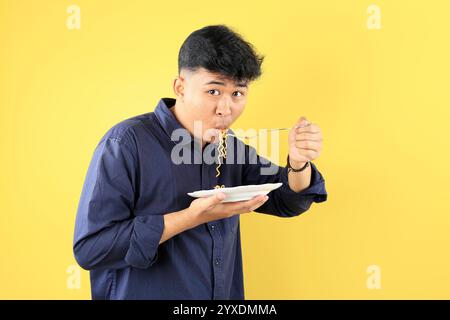 Giovane studente indonesiano che mangia spaghetti istantanei utilizzando forchetta in acciaio inox. Su sfondo giallo Foto Stock