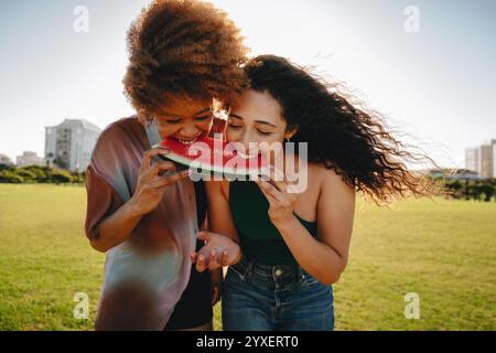Due giovani donne condividono una fetta di anguria in un parco cittadino in una giornata estiva, trasudando gioia e divertimento. Foto Stock