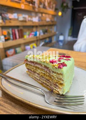 Questa immagine presenta una deliziosa fetta di torta al pistacchio a strati adornata con una liscia glassa verde, pistacchi schiacciati e vivaci petali di rosa commestibili Foto Stock