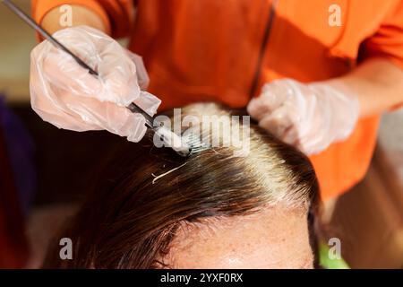 Parrucchiere che indossa guanti che applicano tintura ai capelli di un cliente, coprendo radici grigie in un parrucchiere Foto Stock
