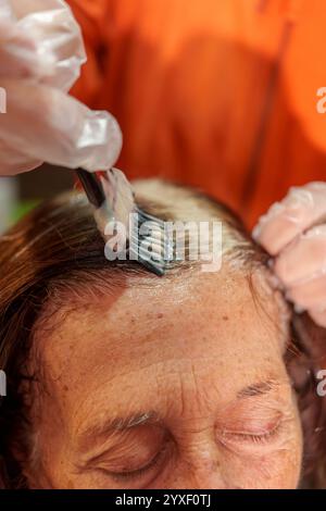 Donna anziana con gli occhi chiusi che si copre i capelli grigi da un parrucchiere che indossa guanti e applica colorante Foto Stock