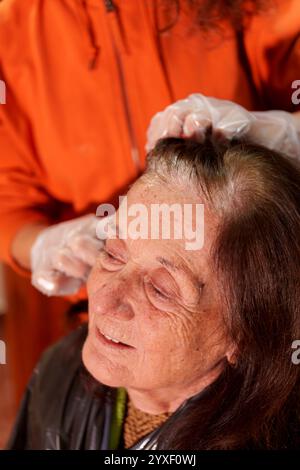 Parrucchiere che indossa guanti, applica tinture per capelli a una donna anziana con capelli grigi, creando un nuovo look vivace nel comfort di casa Foto Stock