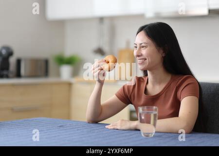 Donna asiatica che mangia ciambella contemplando in cucina a casa Foto Stock