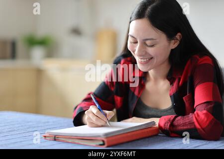 Felice studente asiatico che scrive appunti su un taccuino in cucina a casa Foto Stock