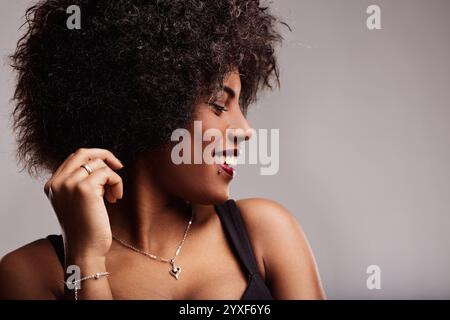 Donna latinoamericana sorridente con capelli ricci, toccandola e guardando via, irradia libertà e fiducia in un elegante ritratto da studio Foto Stock