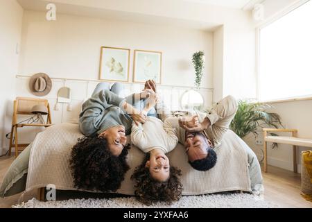 Una famiglia allegra trascorre del tempo insieme a casa giocando a testa in giù su un letto. I genitori e i bambini condividono le risate in una camera accogliente. Foto Stock