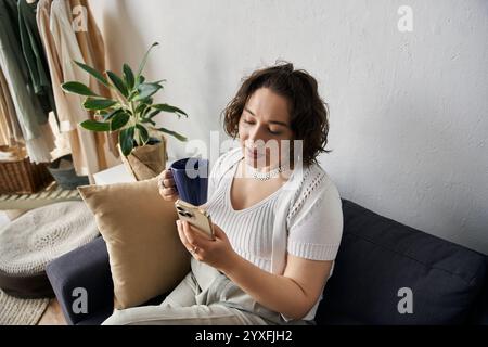 Una giovane donna con i capelli ricci si diverte a bere mentre si controlla il telefono in uno spazio accogliente. Foto Stock