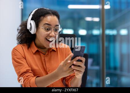 Una donna allegra che indossa le cuffie reagisce con entusiasmo mentre usa il suo smartphone al lavoro. L'ambiente d'ufficio suggerisce un ambiente professionale, ricevendo buone notizie. Foto Stock