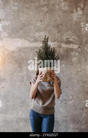 La donna si trova contro un muro grigio di cemento e tiene un albero di Natale davanti al viso Foto Stock