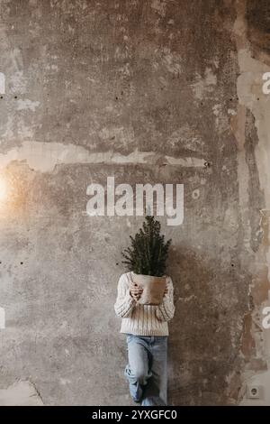 La ragazza sta contro un muro di cemento grigio e tiene un albero di Natale davanti al viso Foto Stock