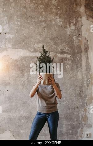 La donna si trova contro un muro grigio di cemento e tiene un albero di Natale davanti al viso Foto Stock