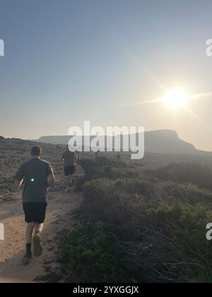 Questa immagine cattura un gruppo di individui che corrono lungo un aspro sentiero costiero all'alba ad Ayia Napa, Famagosta, Cipro. Il sole nascente getta un caldo Foto Stock
