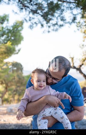 Un padre tiene il suo bambino sorridente in un ambiente tranquillo all'aperto. L'immagine cattura un momento gioioso di legame e felicità circondato dalla natura Foto Stock