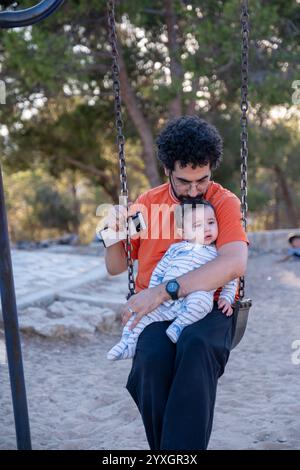 Un momento sincero catturato nel parco mentre padre e bambino condividono una pausa tranquilla sull'altalena, incarnando le semplici gioie del tempo trascorso in famiglia all'aperto. Foto Stock