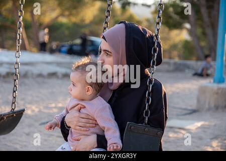 Un momento emozionante che cattura una madre musulmana in abaya tradizionale e hijab in stile arabo, condividendo la gioia del parco giochi con il suo bambino, mescolando va culturale Foto Stock