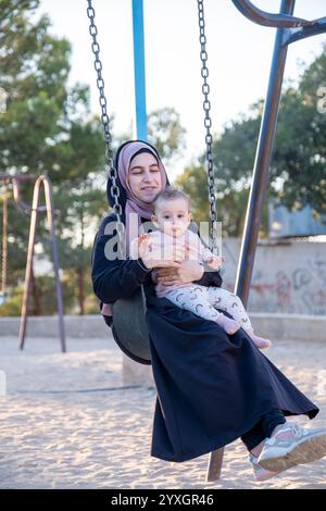 Un momento emozionante che cattura una madre musulmana in abaya tradizionale e hijab in stile arabo, condividendo la gioia del parco giochi con il suo bambino, mescolando va culturale Foto Stock