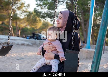 Un momento emozionante che cattura una madre musulmana in abaya tradizionale e hijab in stile arabo, condividendo la gioia del parco giochi con il suo bambino, mescolando va culturale Foto Stock