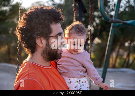 Un padre amorevole tiene il suo bambino mentre si diverte insieme in un parco soleggiato. Il momento cattura il calore, la connessione e la gioia della famiglia bondi Foto Stock