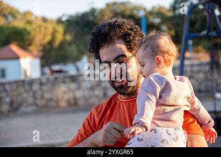 Un padre amorevole tiene il suo bambino mentre si diverte insieme in un parco soleggiato. Il momento cattura il calore, la connessione e la gioia della famiglia bondi Foto Stock