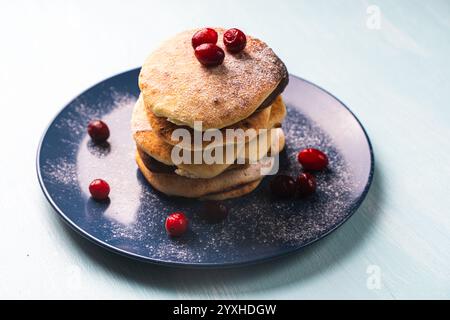 Le frittelle di formaggio con mirtilli rossi sono impilate su un piatto blu su un tavolo azzurro, cosparse di zucchero a velo. Foto di alta qualità Foto Stock