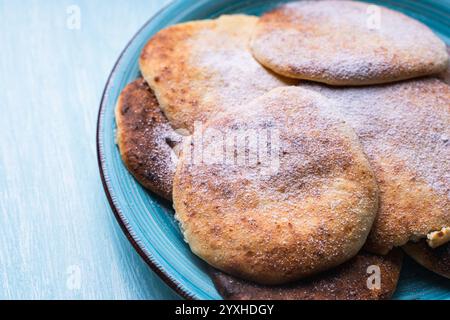 Le frittelle di formaggio con mirtilli rossi sono impilate su un piatto blu su un tavolo azzurro, cosparse di zucchero a velo. Foto di alta qualità Foto Stock