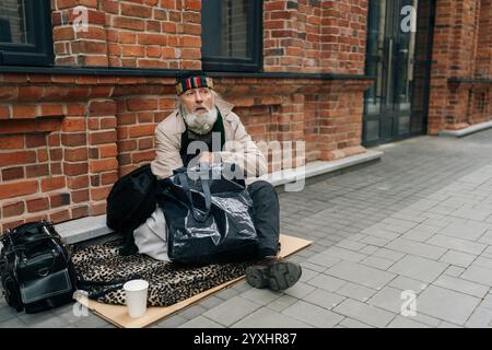 Ritratto di un vecchio sconsiderato con la lunga barba bianca seduto su una strada urbana con pochi effetti personali, guardando verso l'alto con speranza, sperimentando senzatetto e. Foto Stock